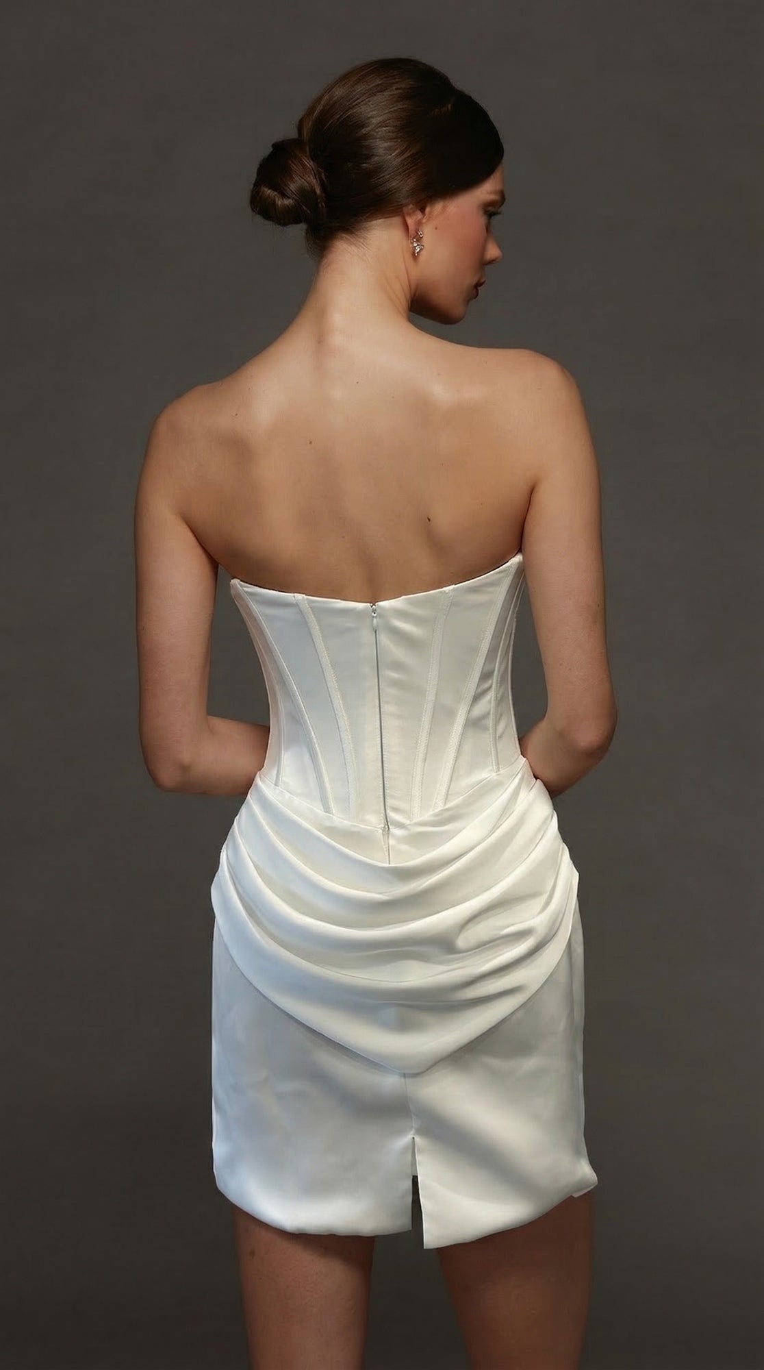 A woman with dark hair in a low bun is seen from the back against a plain dark background, wearing the Alyce 70090 short white party dress featuring a strapless satin design with a fitted corset bodice and draped skirt.